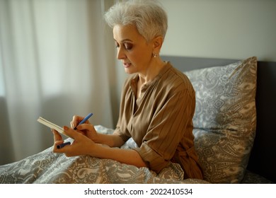 Portrait Of Busy Modern 50 Year-old-middle Aged Woman In Brown Shirt Writing In Copybook, Making Plans, Having Deep-in-thoughts Face Expression And Concentrated Look, Sitting On Bed