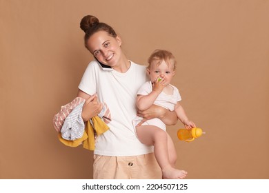 Portrait Of Busy Mother Holding Baby Daughter And Clothing Isolated Over Brown Background, Female Wearing White T Shirt Talking On Mobile Phone, Expressing Positive Emotions.