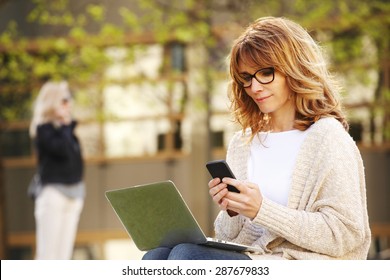 Portrait of busy sales woman sitting at business park and using her laptop while writing text on mobile. Businesswoman working online. 