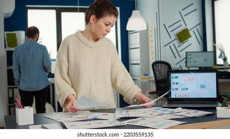 Portrait Of Busy Startup Employee Holding Papers With Business Charts Comparing Smiling At Camera Standing At Desk With Laptop. Confident Woman Analyzing Financial Data In Small Company Office.