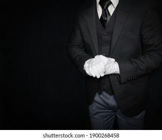Portrait Of Butler Or Servant In Dark Suit And White Gloves Ready To Help On Black Background. Service Industry. Professional Hospitality And Courtesy. Formal White Glove Service.