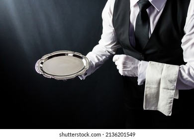 Portrait of Butler or Waiter in Black Vest and White Gloves Holding Serving Tray on Black Background. Concept of Service Industry and Professional Courtesy.