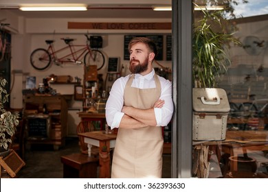 Portrait Of A Cafe Owner Standing In The Doorway Of His Coffee Shop. Young Waiter Standing With His Arms Crossed At The Door Of A Restaurant And Looking Away.