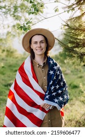 Portrait Of Calm Beautiful Young Caucasian Woman 20s In Beige Hat Wrapped In American Flag. Hipster Girl Smiling And Looking At Camera On Background Forest. July 4th Forth Independence Day Concept