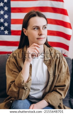 Portrait of calm beautiful young girl 20s old years sitting on comfortable couch at home on background USA flag. 4th of July, Independence day celebration. Patriotic holiday, democracy concept