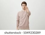 Portrait of calm serious young man with bristle stretching hand towards camera with stop or hold gesture, standing against gray background. You shall not pass. Officer forbids to enter area