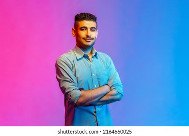 Portrait of calm smiling man in shirt standing with crossed arms, looking at camera with confident facial expression. Indoor studio shot isolated on colorful neon light background.