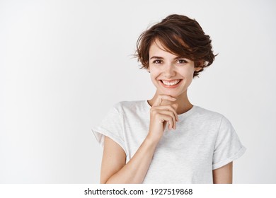 Portrait of candid happy woman smiling and touching chin, looking pensive, standing against white background.