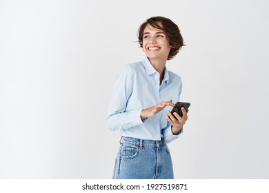 Portrait Of Candid Young Woman Using Mobile Phone And Smiling, Looking At Empty Space For Logo, White Background.