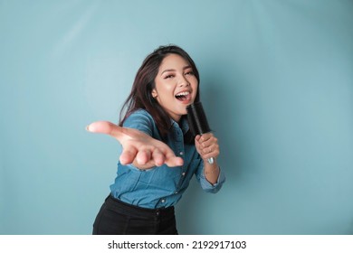 Portrait of carefree Asian woman, having fun karaoke, singing in microphone while standing over blue background