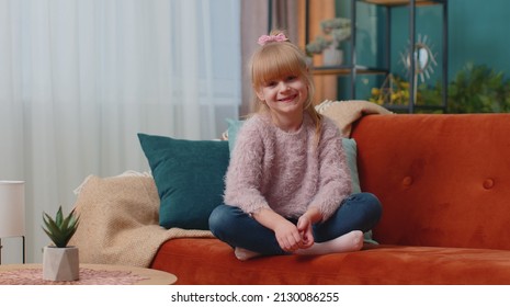 Portrait of carefree Caucasian little adorable happy child girl sitting on sofa in living room looking at camera and smiling in positive mood. Happy childhood. Small cute teen school kid at home alone