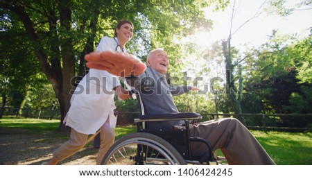 Portrait of carefree and happy young woman social worker or nurse  and senior man in a wheelchair having fun to run in a green park on a sunny day.