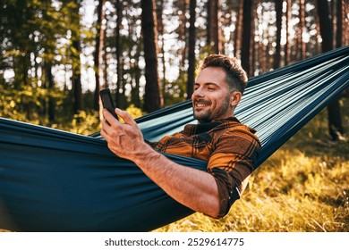 Portrait of carefree man using mobile phone in hammock during afternoon in forest - Powered by Shutterstock