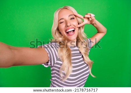 Portrait of carefree positive girl make selfie arm fingers demonstrate v-sign eye wink isolated on green color background