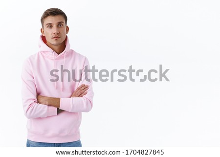 Portrait of carefree, relaxed handsome blond man in pink hoodie, cross arms over chest and look camera serious, getting straight to business, listening to information, standing white background