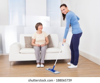 Portrait Of Caretaker Cleaning Floor With Mop While Senior Woman Sitting On Sofa At Nursing Home