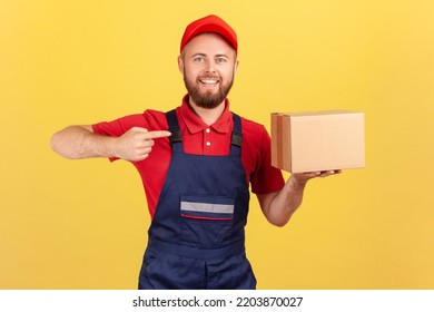 Portrait Of Cargo Transportation, Delivery Service. Friendly Courier Man In Uniform Pointing At Cardboard Box, Delivering Parcel To Client. Indoor Studio Shot Isolated On Yellow Background.