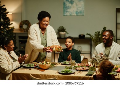 Portrait of caring African-American grandmother bringing food to table while celebrating Thanksgiving with big happy family