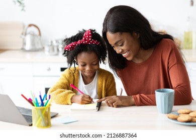 Portrait Of Caring Black Mom Helping Her Little Daughter With Homework, African American Mother And Cute Female Child Sitting At Table In Kitchen, Girl Writing With Pen In Notepad, Closeup