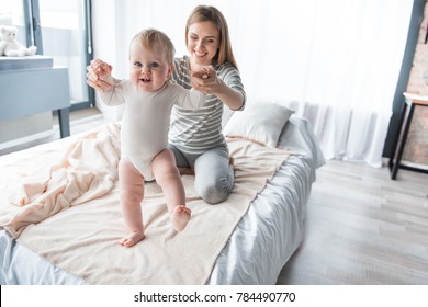 Portrait of caring mom sitting on bed and teaching her little kid walking. Focus on baby. Copy space in right side