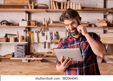 Portrait Of A Carpenter Smiling At The Camera While Listening On His Mobile Phone, Standing In His Woodwork Studio And Holding A Digital Tablet