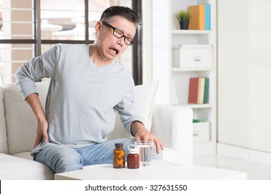 Portrait of casual 50s mature Asian man back pain, pressing on hip with painful expression, sitting on sofa at home, medicines and water on table.