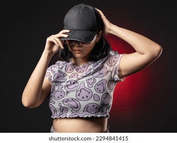 A portrait of a casual fashion style Asian Girl wearing a black cap posing with a Hip hop style. Isolated on Black Background with a red spotlight - Powered by Shutterstock