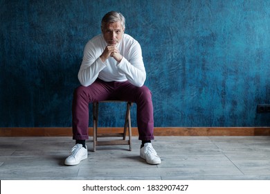 Portrait Of A Casual Grey-haired Mature Handsome Man Sitting On A Chair On Dark Blue Background