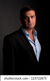 Portrait Of A Casual Middle Aged Senior Executive Taken Using Low Key Lighting.  He Has Brown Hair, Brown Eyes, And Is Wearing A Grey Color Suit, And Light Blue Shirt, Albeit No Tie.