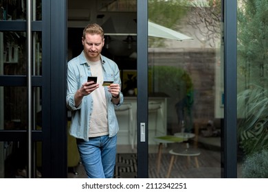 Portrait of casual relaxed man using phone for online payment on doorstep of his home, shopping online from home leaning on door frame using credit card, casual entrepreneur doing business from home