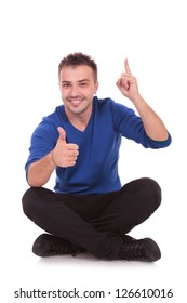 Portrait Of A Casual Young Man Sitting On The Floor, Legs Crossed, Giving The Thumbs Up And Pointing Upwards, All This With A Big Smile On His Face.