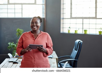 Portrait of a casually dressed young African businesswoman smiling and using a digital tablet while standing alone in a modern office - Powered by Shutterstock