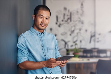 Portrait Of A Casually Dressed Young Asian Designer Smiling And Using A Digital Tablet While Leaning Against A Gray Wall In A Modern Office