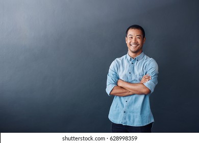 Portrait Of A Casually Dressed Young Asian Designer Standing Confidently With His Arms Crossed Against A Gray Wall In An Office