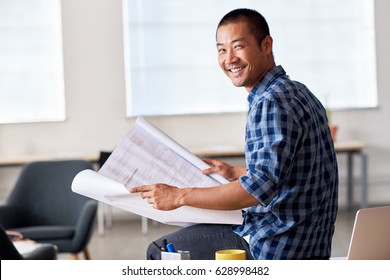 Portrait Of A Casually Dressed Young Asian Architect Smiling Confidently While Reading Blueprints In A Modern Office Reading Blueprints