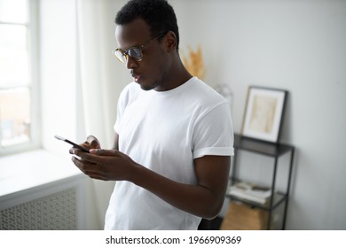 Portrait Of Casually Dressed Young Dark Skinned Man With Bristle Posing At Home Holding Cell Phone, Typing Text Message To Friends, Making Plans On Weekend. Black Guy Shopping Online Using Mobile