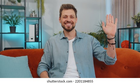 Portrait Of Caucasian Adult Man Smiling Friendly At Camera And Waving Hands Gesturing Hello, Hi, Greeting Or Goodbye, Welcoming With Hospitable Expression. Young Guy Indoor At Home In Room On Couch