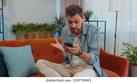 Portrait Of Caucasian Adult Man Use Smartphone Typing Browsing, Loses Becoming Surprised Sudden Lottery Results, Bad News, Fortune Loss, Fail. Young Guy Sitting On Orange Couch At Home Living Room