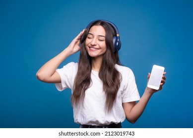 Portrait of a Caucasian or Arab young woman listening to her favorite music on wireless headphones from her mobile phone wearing a white casual t-shirt isolated on a blue studio background. - Powered by Shutterstock