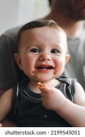 Portrait Of An Caucasian Baby Girl Sitting In Her Father's Arms With A Happy Smile, Teeth Out And A Face Dirty From Pizza Sitting In The Kitchen At A Round Table Against The Window, Close-up Side View