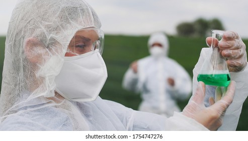 Portrait Of Caucasian Beautiful Young Female Biologist Researcher In Protective Costume, Mask And Goggles Holding Flask With Chemicals And Smiling To Camera In Field.