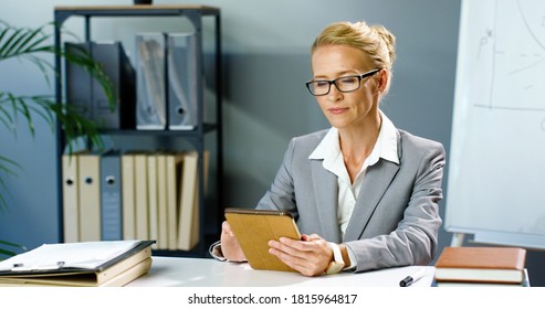 Portrait Of Caucasian Businesswoman In Glasses Sitting In Office And Using Tablet Device, Smiling To Camera. Female In Business Style Tapping And Scrolling On Gadget Computer At Table In Cabinet.