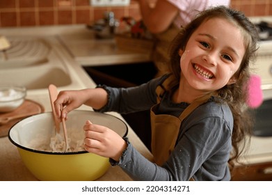 Portrait of a Caucasian cheerful and mischievous little confectioner, an adorable baby girl in a chef's apron, smiling cheerfully, looking at the camera and kneading dough to bake a festive cherry pie - Powered by Shutterstock