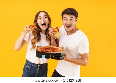 Portrait of caucasian couple man and woman in basic t-shirts smiling while eating pizza from box isolated over yellow background
