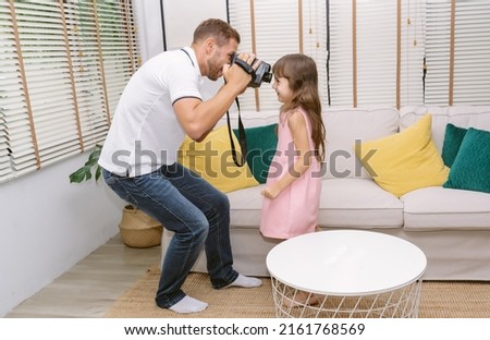 Portrait of Caucasian Father taking video of his little daughter with old retro video camcorder camera in living room at home. Family holiday and togetherness enjoying free time.