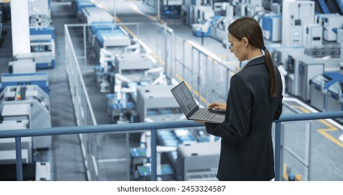 Portrait of a Caucasian Female Engineer Using Laptop Computer and Looking Around a Factory Facility with Robotic Arms Producing Modern Electronic Components for Tech Industries - Powered by Shutterstock