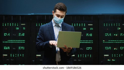 Portrait of Caucasian handsome male security engineer in mask in datacenter checking security of processors with digital information working in data storage and typing on laptop. Technologies concept