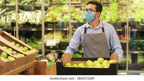 Portrait Of Caucasian Handsome Man Worker In Mask And Apron Walking In Supermarket With Shopping Cart With Fruits Indoor. Male Young Food Store Assistant In Glasses Working In Retail. Job Concept