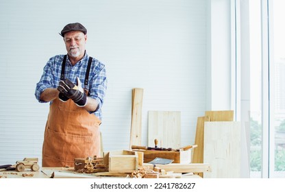 Portrait Caucasian happy old male woodworker or carpenter smiling, wearing shirt with apron, vintage hat, safety gloves for creating DIY wooden furniture as hobby after retirement - Powered by Shutterstock