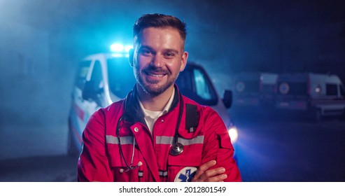 Portrait Of Caucasian Happy Young Male Paramedic In Red Uniform Smiling To Camera And Standing Outdoor. Ambulance Car On Background. Attractive Cherful Male Doctor At Night Shift. Call 911